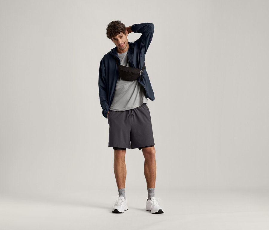 Mężczyzna pozuje z ręką na głowie, ma na sobie jasnoszarą koszulkę sportową, ciemną kurtkę, szorty i torbę na biodra.