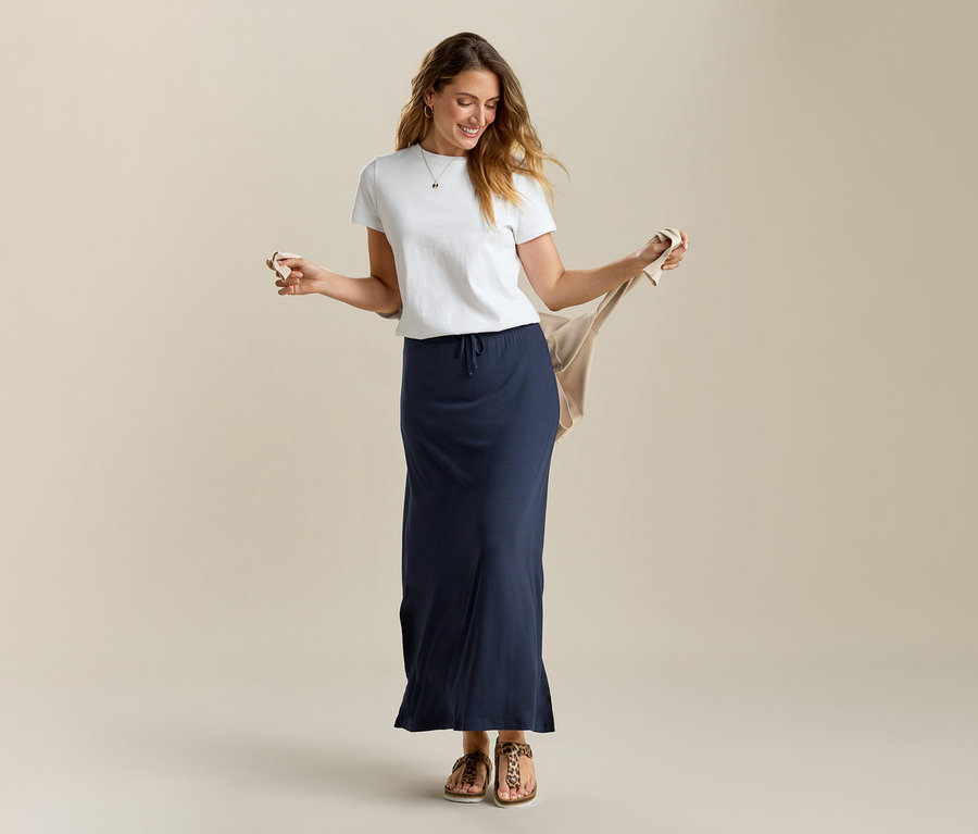 Kobieta trzyma beżowy kardigan, ma na sobie biały T-shirt i długą dżersejową spódnicę w kolorze granatowym.