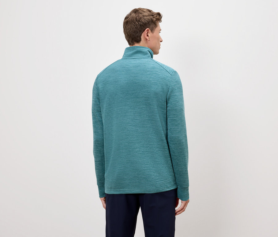 Widok z tyłu mężczyzny o jasnobrązowych włosach w turkusowym pulowerze zapinanym na zamek i granatowych spodniach na kremowym tle.