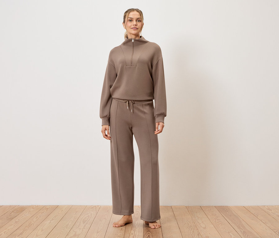 Kobieta stoi boso na drewnianej podłodze, ubrana w brązowy dres.