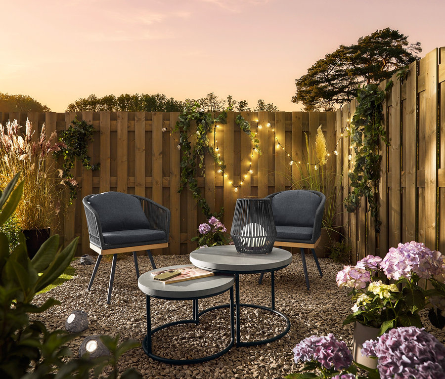 Dwa obrotowe krzesła ogrodowe z krzyżowym splotem tekstylnym stoją obok 2 stolików ogrodowych z blatem z polikamentu. Mały, solarny lampion ze sznurkiem tekstylnym stoi na stole. Solarne kamienie LED premium leżą w żwirze.