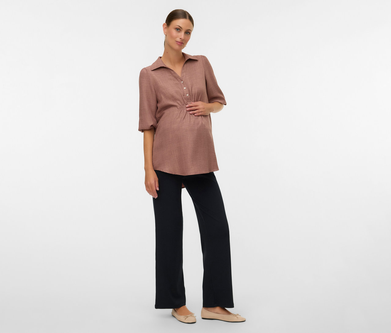 Kobieta w ciąży w brązowej bluzce i luźnych legginsach MAMALICIOUS „MLEMILY” pozuje na białym tle.