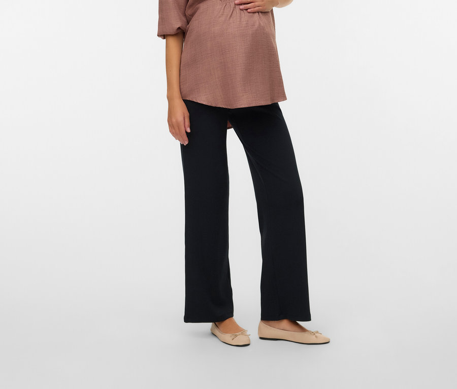 Kobieta w ciąży w jasnobrązowej bluzce i czarnych spodniach, trzymająca ręce na brzuchu.