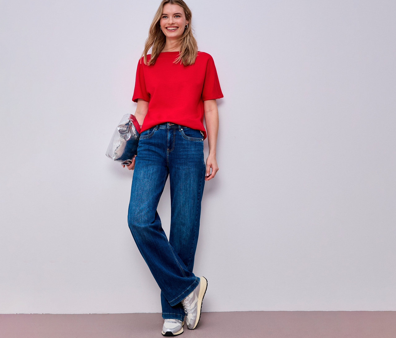 Uśmiechnięta kobieta w czerwonej koszulce Street One, dżinsach i srebrnych butach sportowych trzyma srebrną torbę.