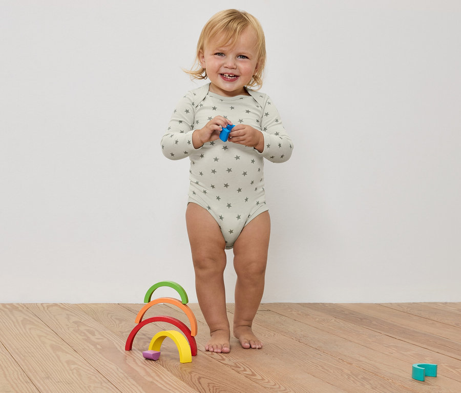 Uśmiechnięte dziecko w body w gwiazdki trzyma zabawkę w dłoniach, przed kolorową drewnianą tęczą na drewnianej podłodze.