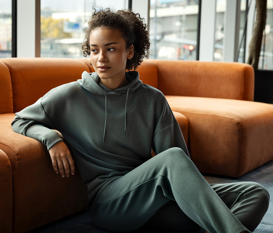 Kobieta siedzi na podłodze, opierając się o pomarańczową sofę, ubrana w zieloną bluzę z kapturem i spodnie dresowe.