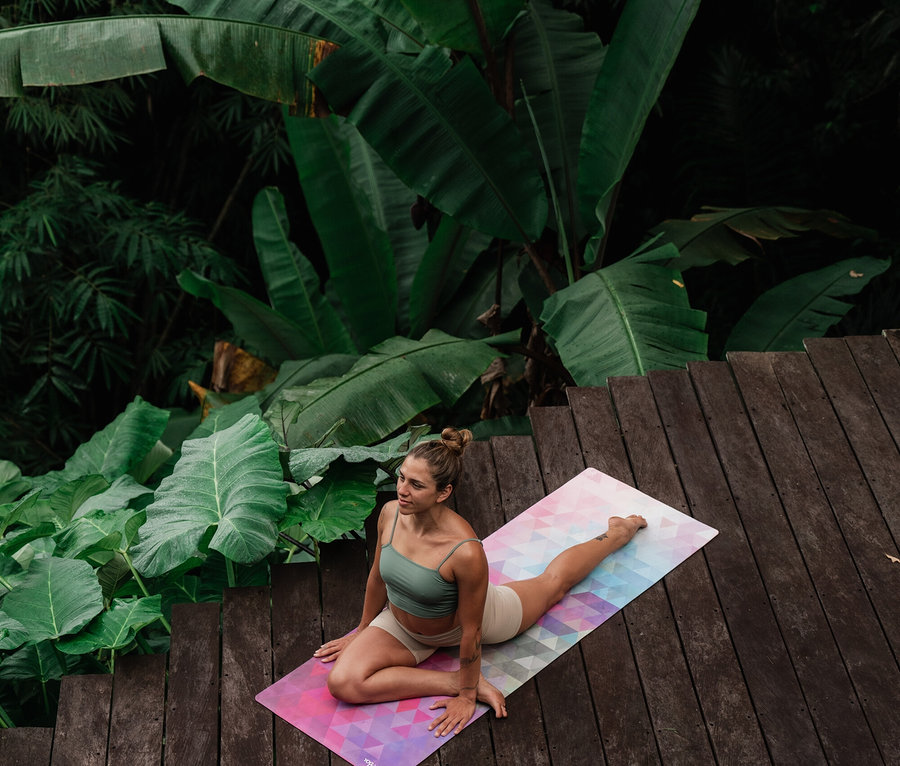 Kobieta rozciąga się na kolorowej macie do jogi na drewnianym tarasie, w tle tropikalna roślinność.