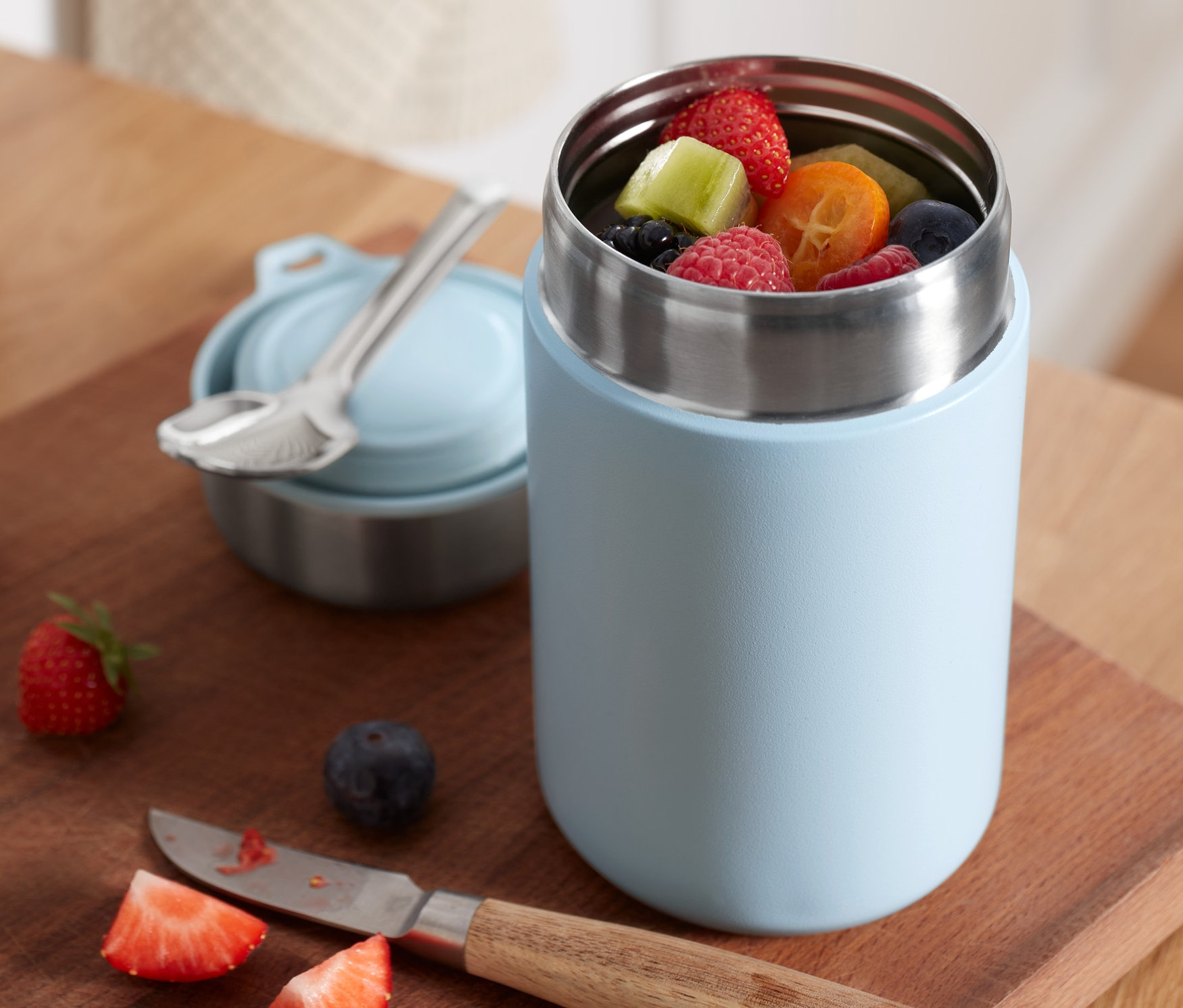 Otwarty pojemnik termiczny wypełniony owocami stoi na drewnianej desce obok pokrywki i łyżki.