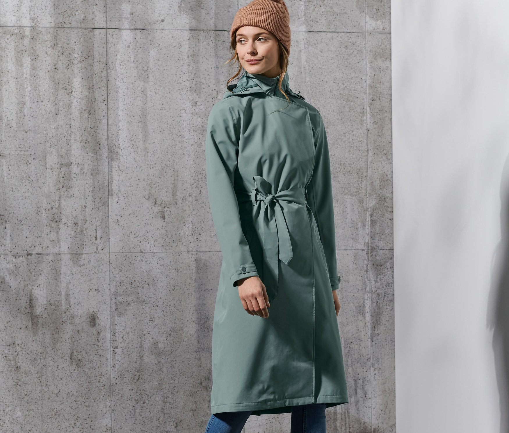 Kobieta pozuje w zielonym płaszczu przeciwdeszczowym, niebieskich dżinsach, beżowych butach i czapce.