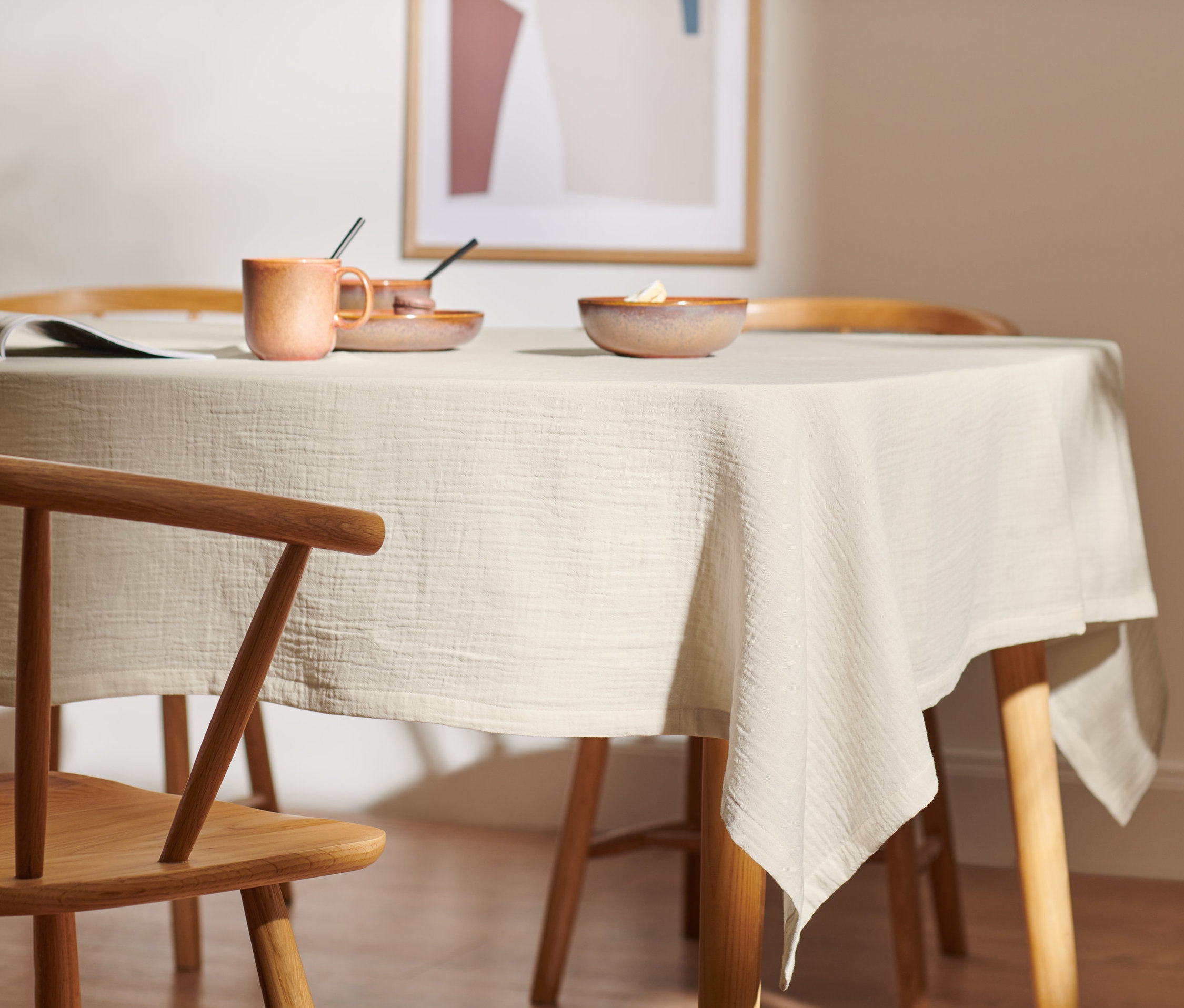 Na stole leży beżowy obrus muślinowy o standardowej wielkości. Na obrusie stoi kubek, talerz i miska.