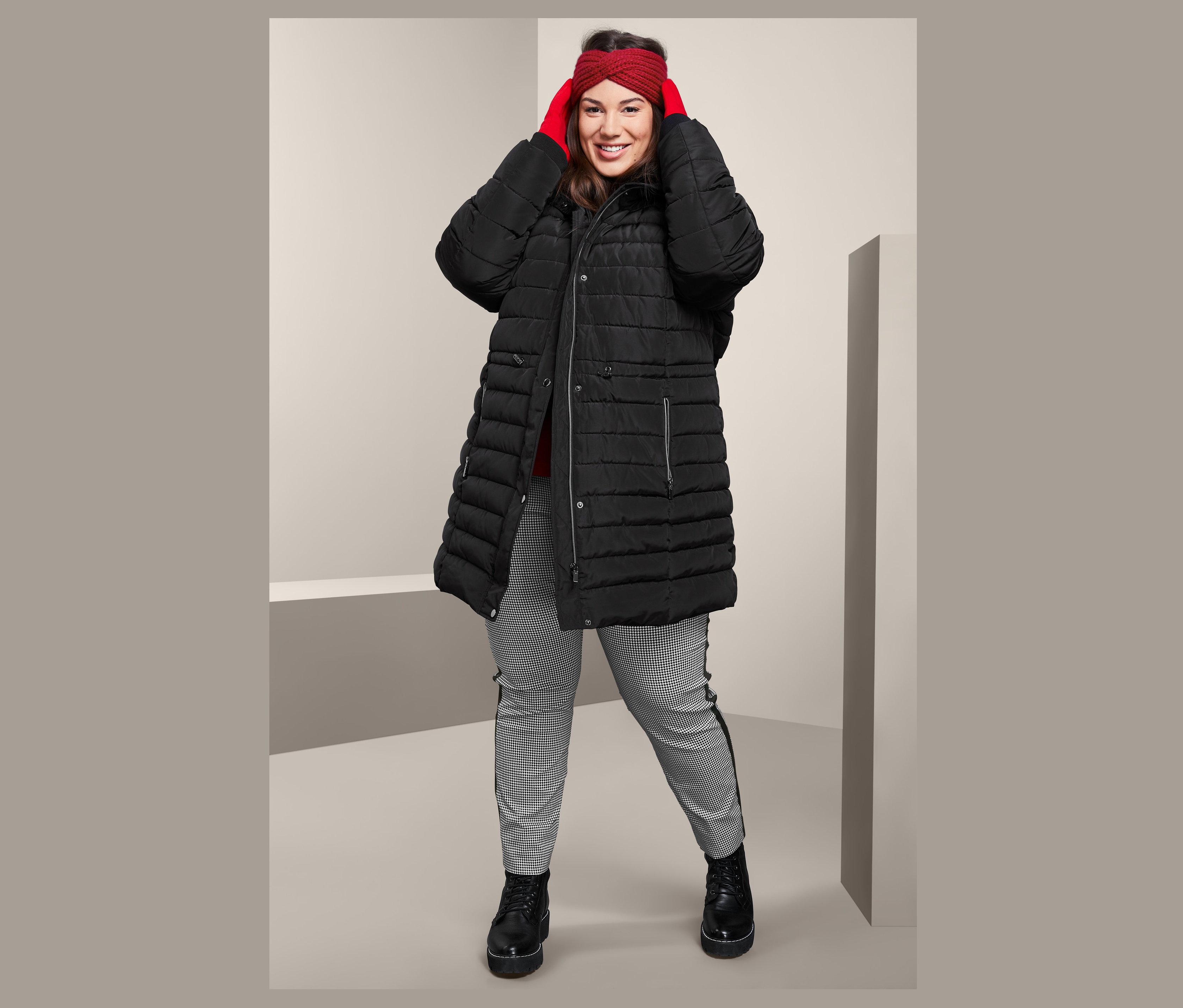 Kobieta poprawia czerwoną opaskę na czoło z dzianiny, ma na sobie czarny damski płaszcz pikowany z kapturem i damskie spodnie ze streczu o długości do kostek.