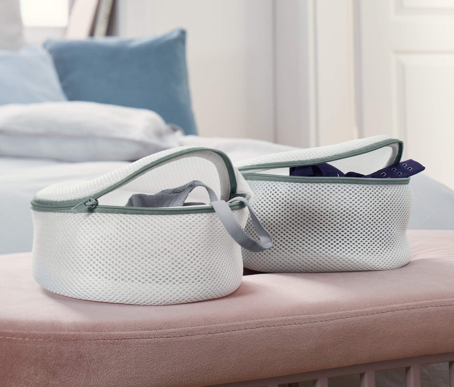 Dwa białe woreczki do prania biustonoszy leżą na różowej ławce przed łóżkiem.