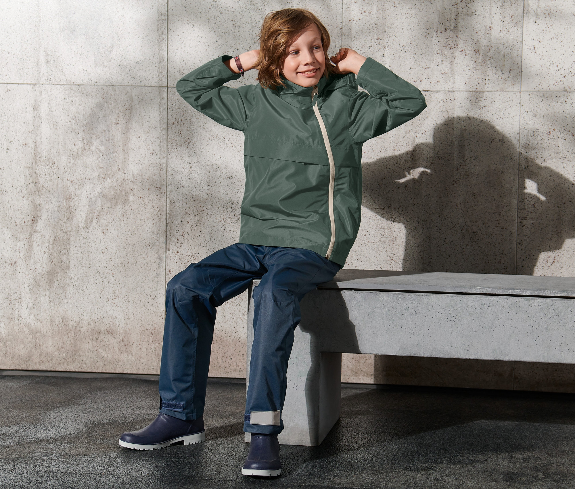 Chłopiec siedzi na ławce i poprawia kaptur kurtki na każdą pogodę.