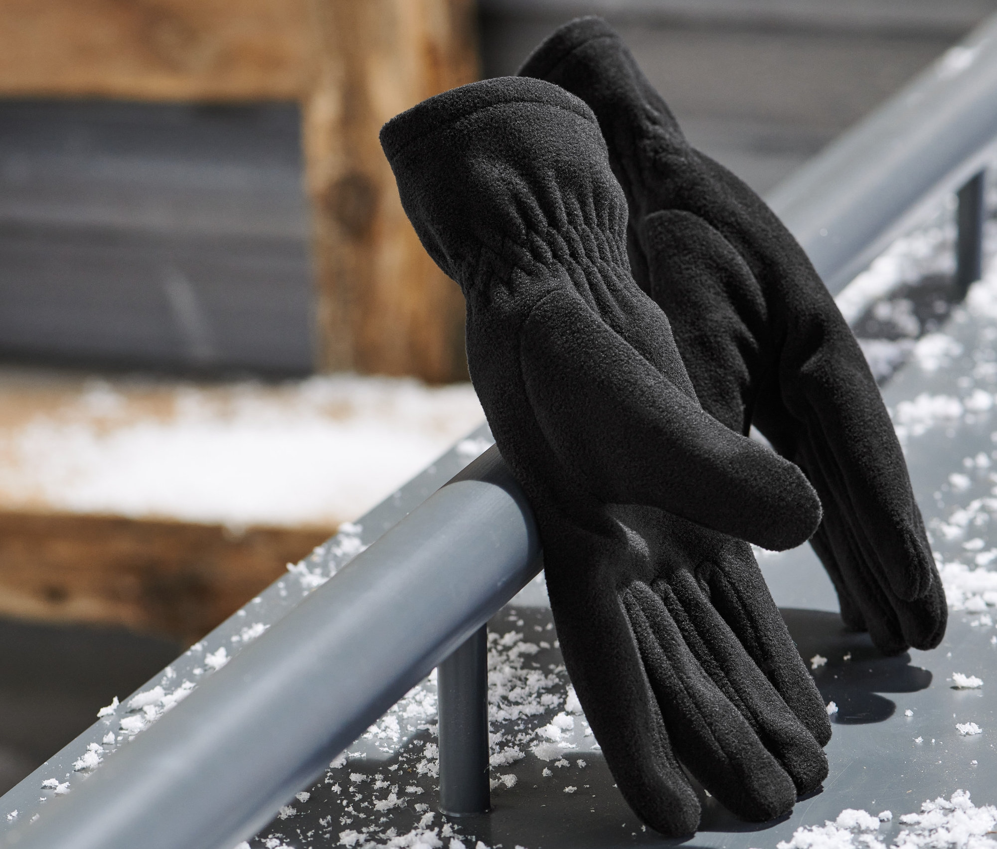 Czarne rękawiczki z mikropolaru leżą na metalowym pręcie. W tle widoczne są gogle narciarskie.