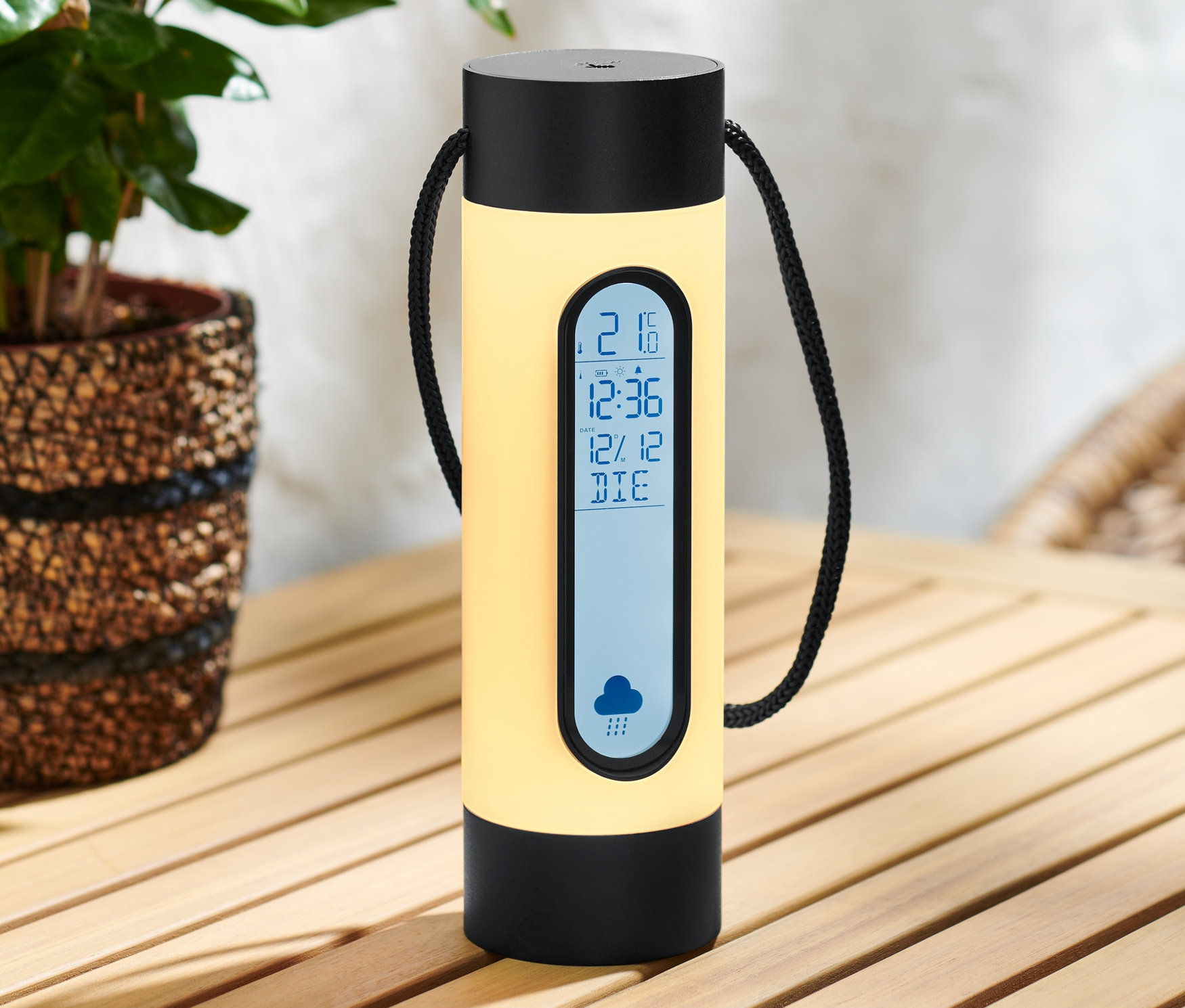 Żółta akumulatorowa stacja pogodowa z lampką stoi na drewnianym stole obok rośliny.