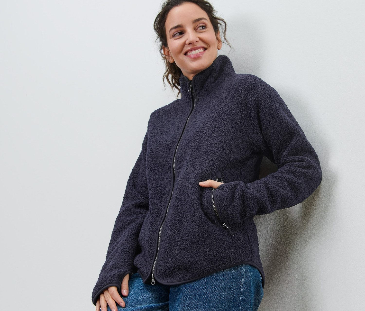 Uśmiechnięta kobieta opiera się o ścianę, ma na sobie granatową bluzę z puszystego polaru i dżinsy.