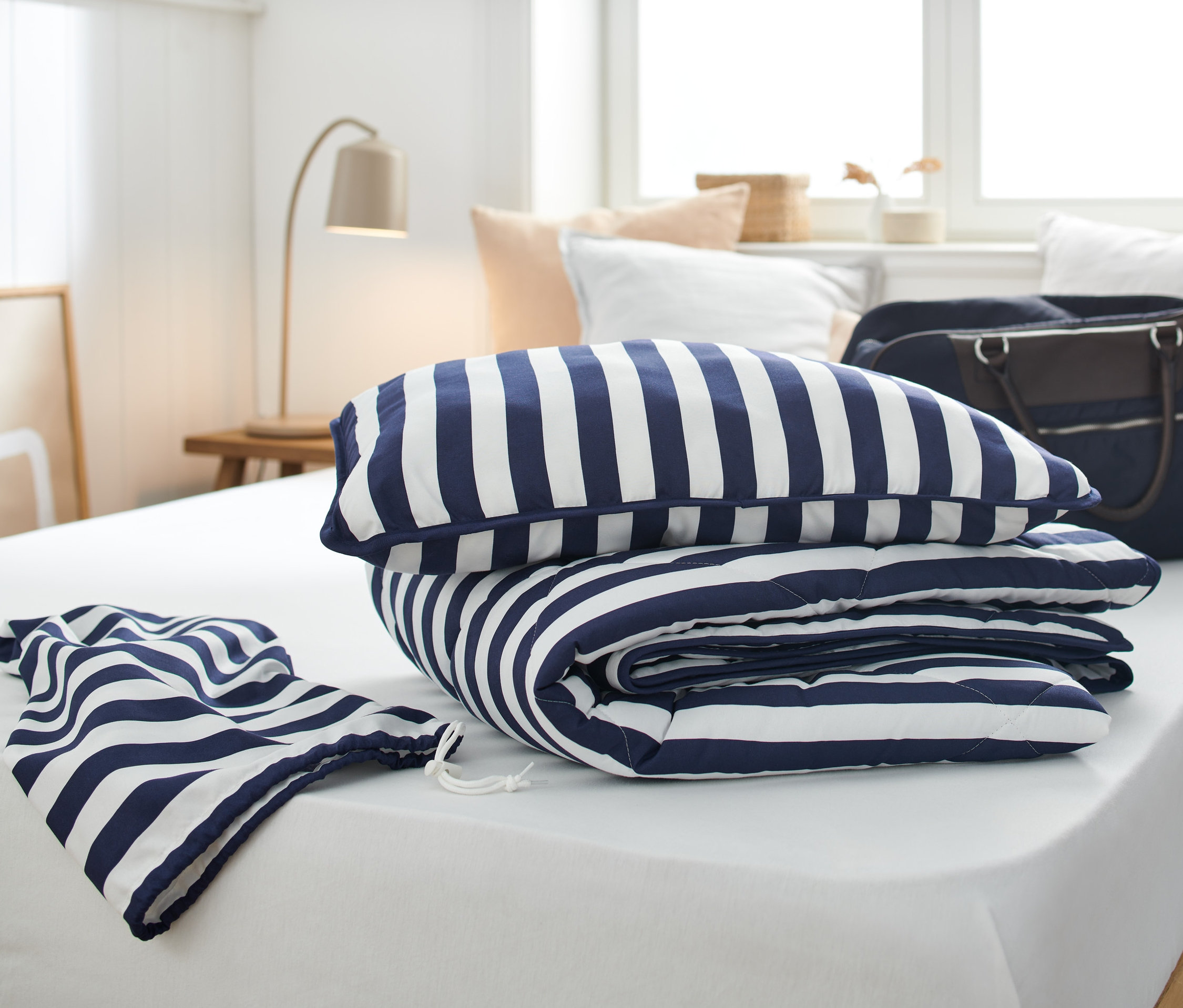 Na białym łóżku leży turystyczny komplet pościeli w niebiesko-białe paski.