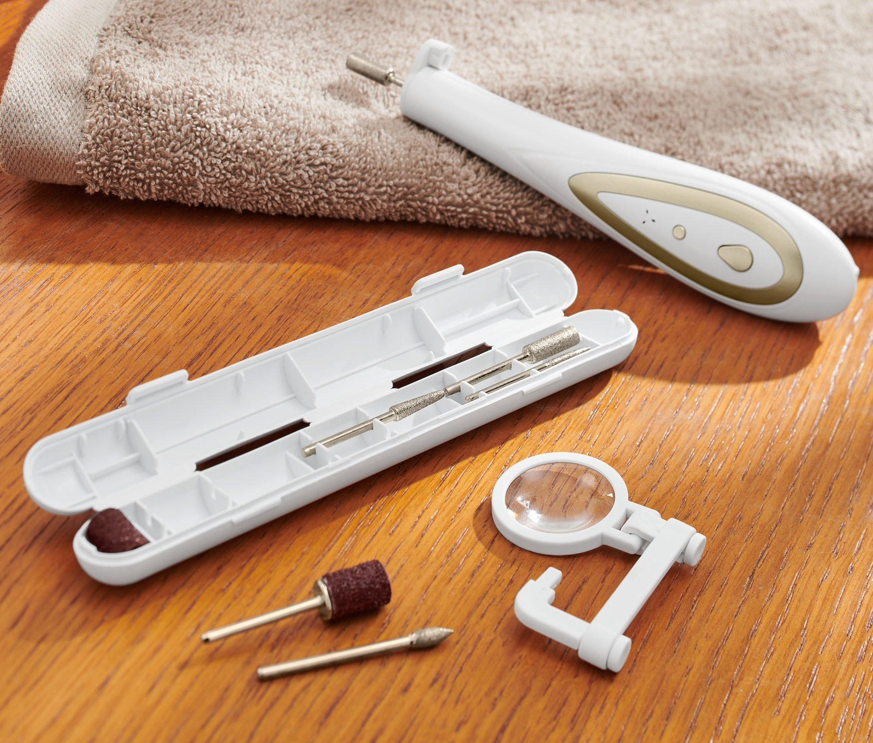 Na drewnianym stole leży urządzenie do manicure i pedicure z otwartym etui na akcesoria, ręcznik i waciki.