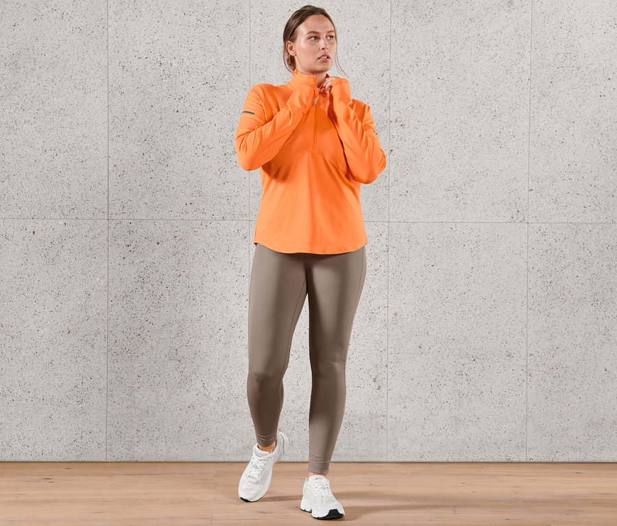 Kobieta ma na sobie neonowopomarańczową termiczną bluzę funkcyjną i legginsy sportowe.