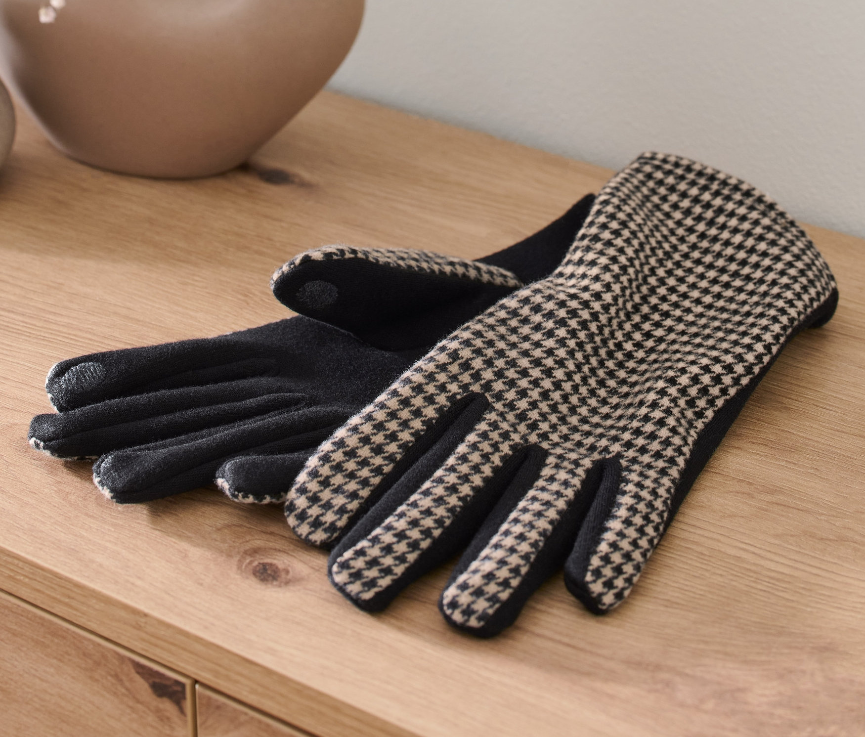 Dwie rękawiczki w pepitkę leżą na drewnianym stole obok wazonu z kwiatami.