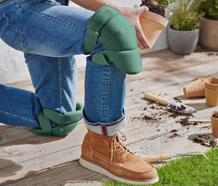 Osoba klęczy w ogrodowych ochraniaczach na kolana, trzymając doniczkę z rośliną.