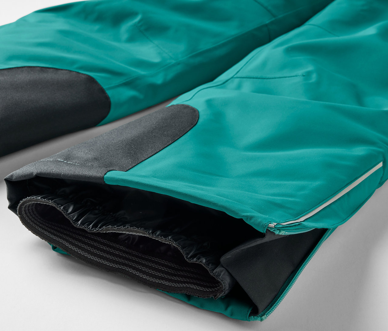 Szczegółowe ujęcie turkusowych spodni z czarnymi łatami na kolanach i rozpiętym brzegiem nogawki, odsłaniającym czarną podszewkę.