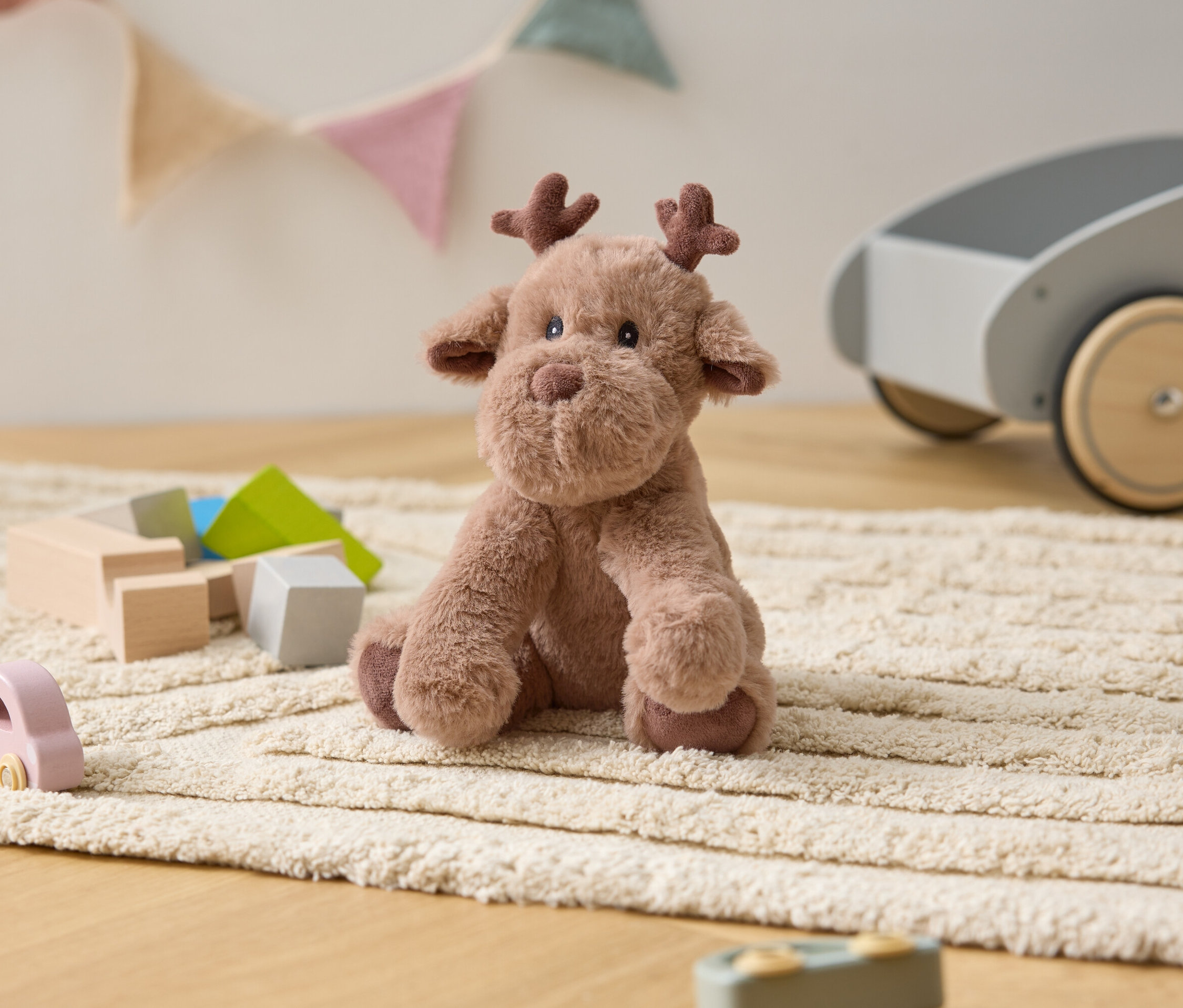 Pluszowy łoś siedzi na kremowym dywanie wśród drewnianych zabawek i szarego wózka.