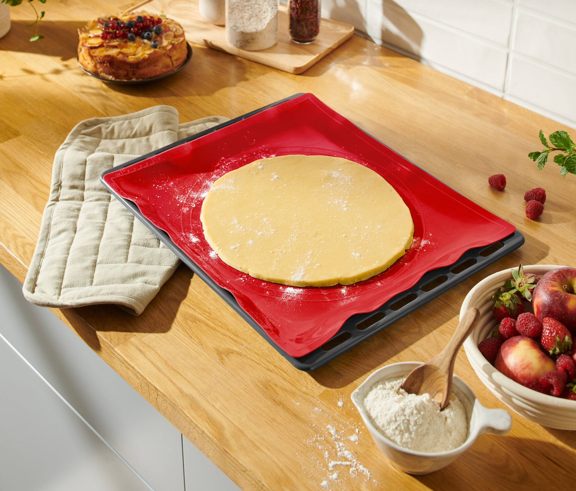 Na blacie leży czerwona mata do pieczenia z rozwałkowanym ciastem, obok leży rękawica kuchenna, misa z owocami i ciasto.