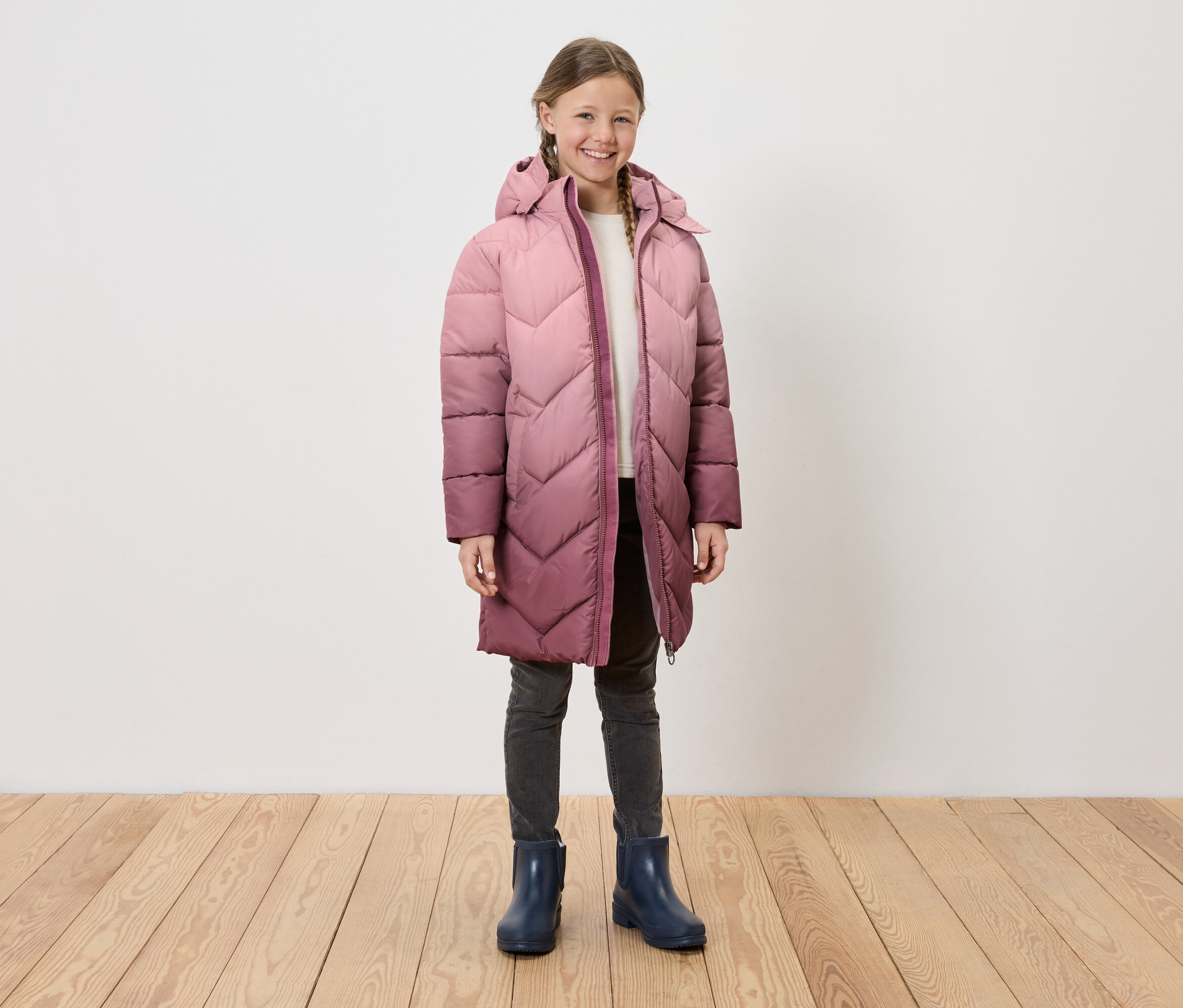 Dziewczyna w długim, pikowanym płaszczu z kapturem, szarych dżinsach i niebieskich kaloszach stoi na drewnianej podłodze przed białą ścianą.