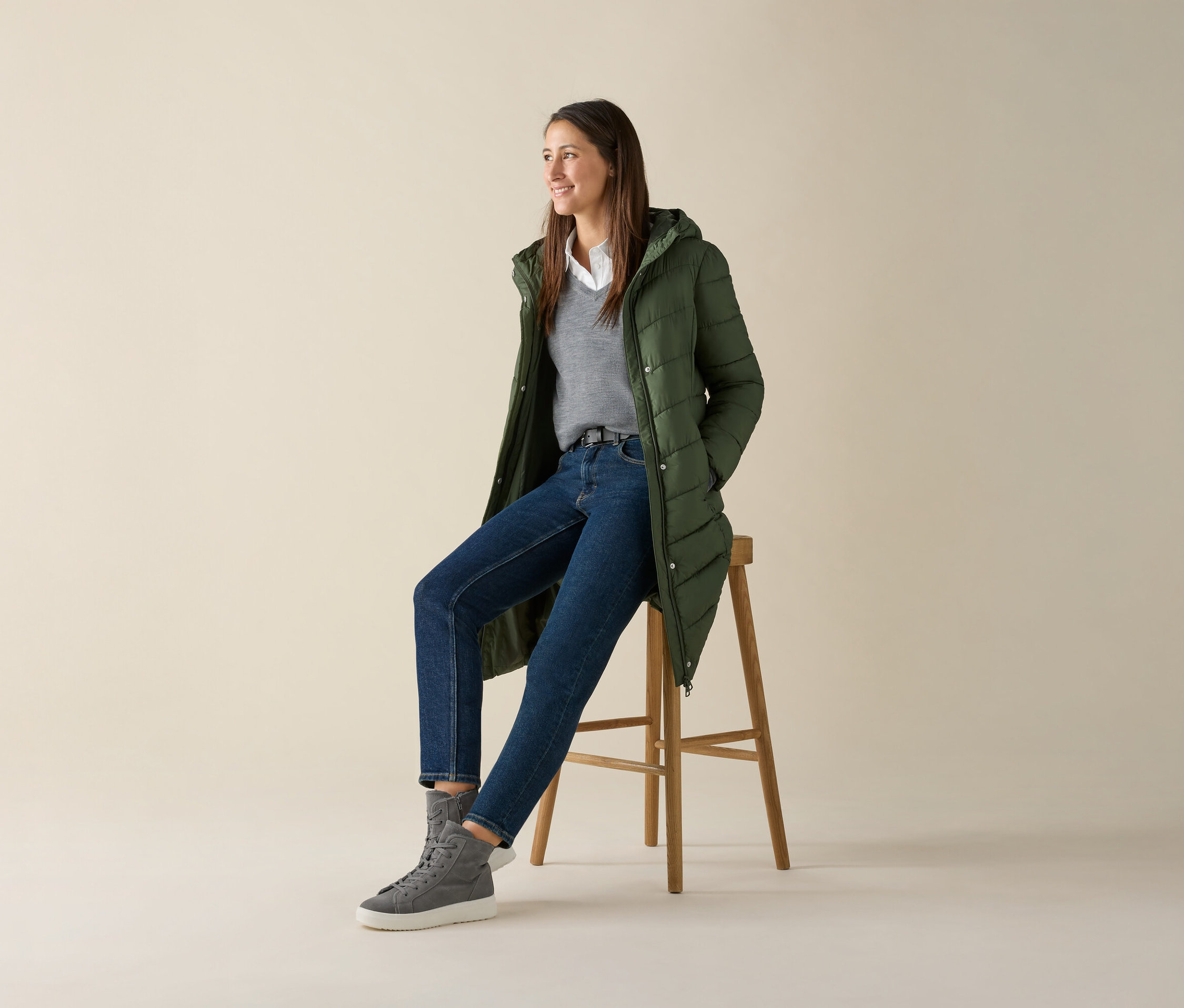 Kobieta siedzi na stołku i ma na sobie szary sweter z cienkiej dzianiny z wełny merino, dżinsy i zieloną kurtkę.