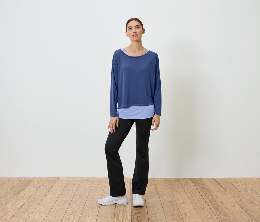 Kobieta stoi na drewnianej podłodze, ubrana w niebieską bluzkę i czarne spodnie.