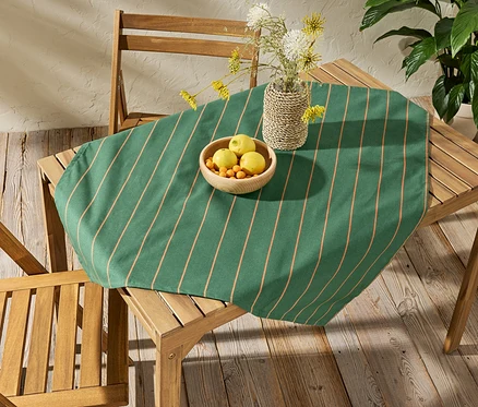 Na drewnianym stole leży obrus ogrodowy w paski o wymiarach ok. 90 x 90 cm. Na obrusie stoi miska z owocami i wazon z kwiatami.