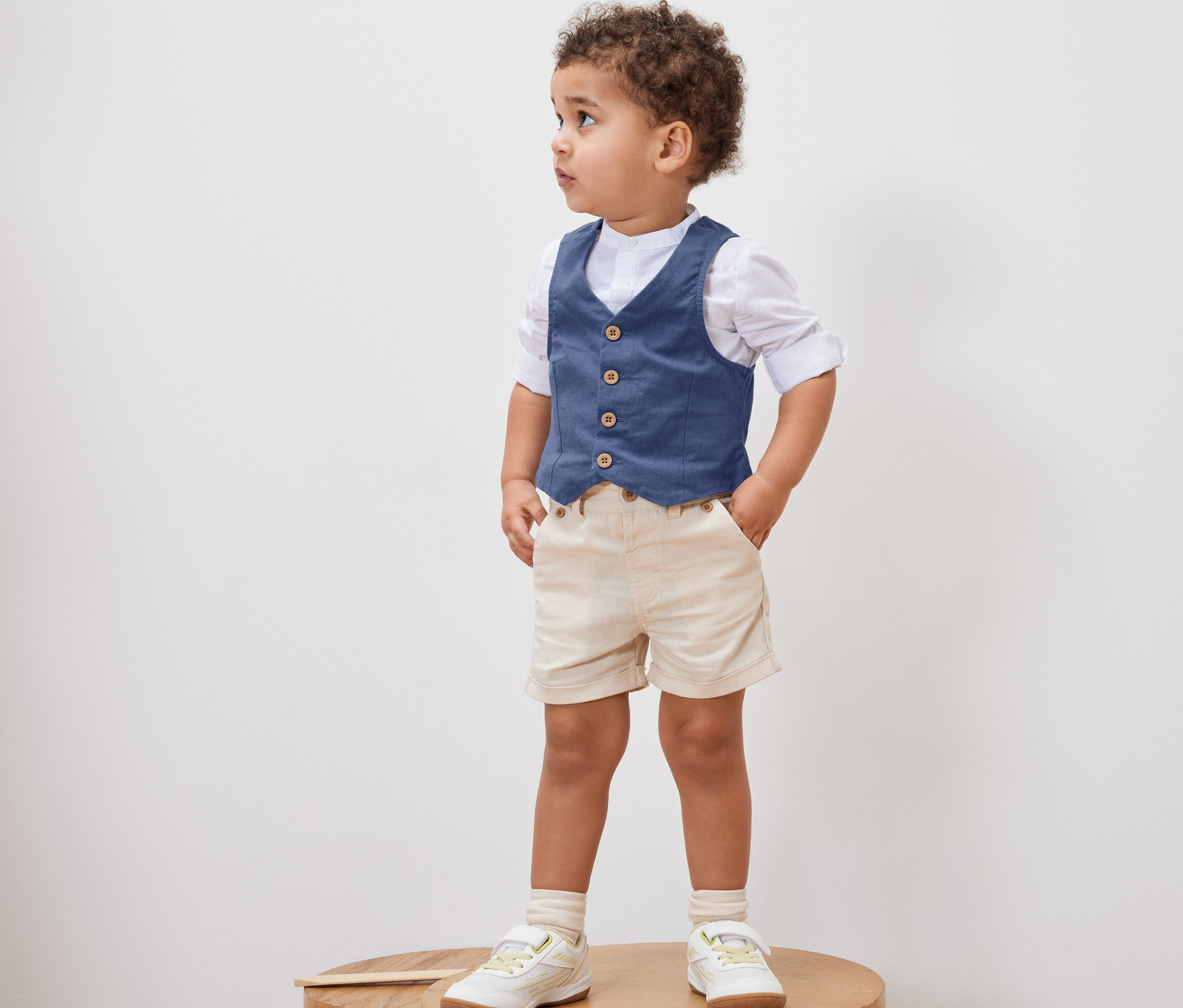Chłopiec stoi na drewnianym stołku, ubrany w Kamizelkę, koszulę, szorty i buty.