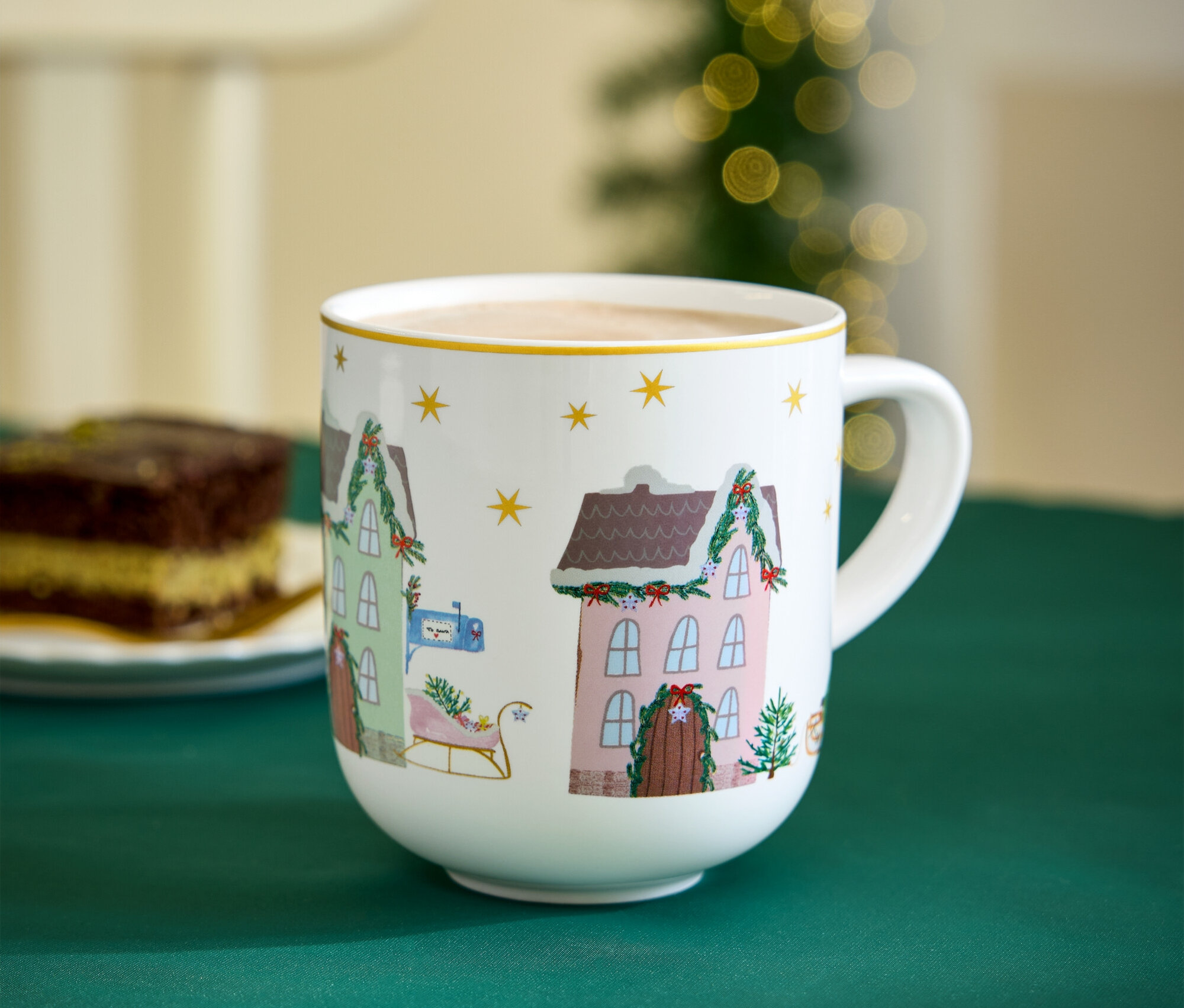 Kubek z motywami świątecznymi stoi na zielonym obrusie obok kawałka ciasta.