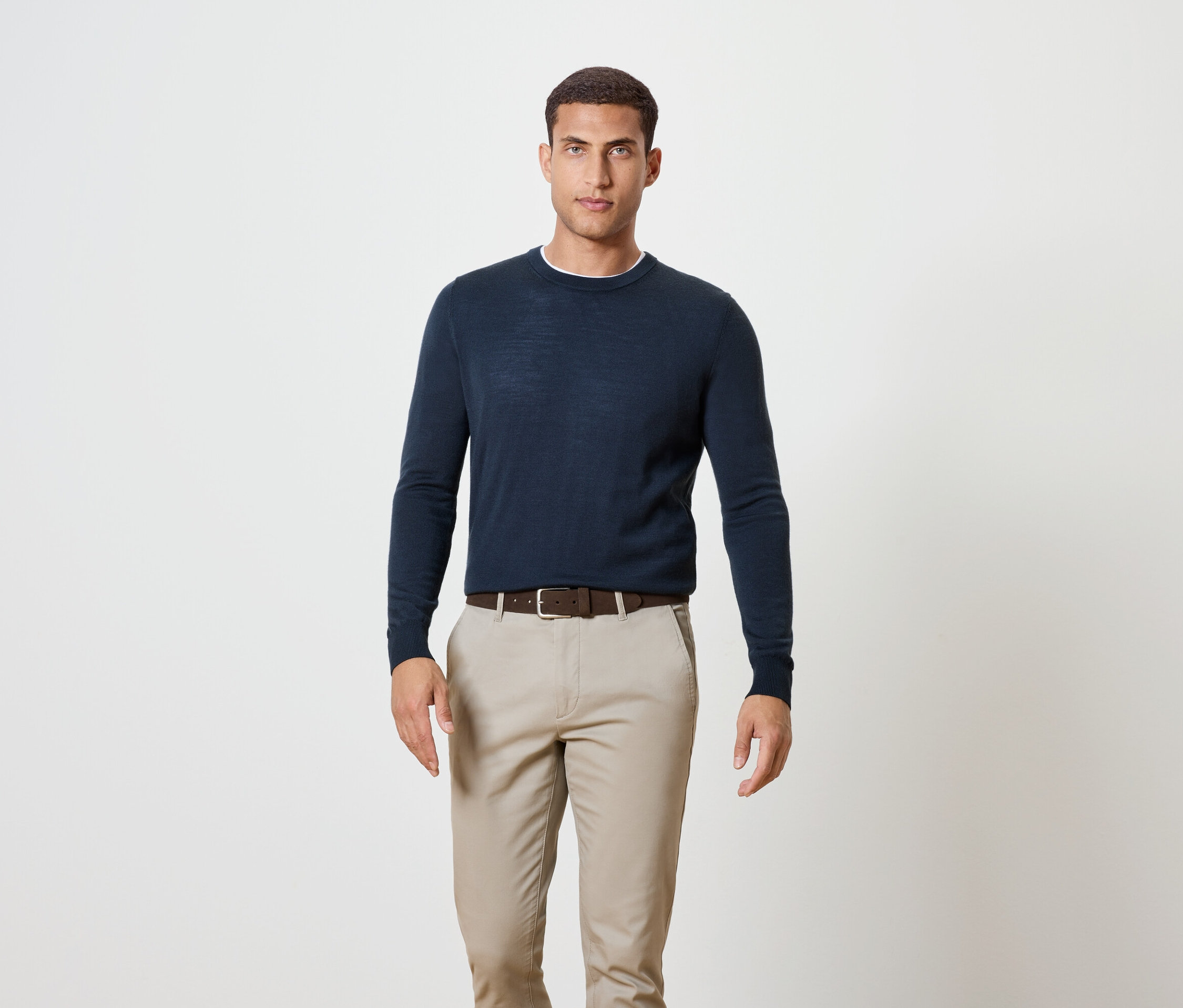 Mężczyzna stoi, ubrany w granatowy sweter, brązowy pasek i spodnie khaki.
