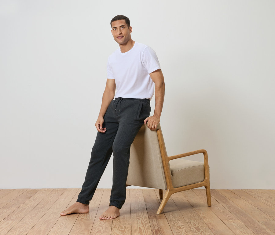 Mężczyzna oparty o krzesło, ubrany w białą koszulkę i szare spodnie dresowe.