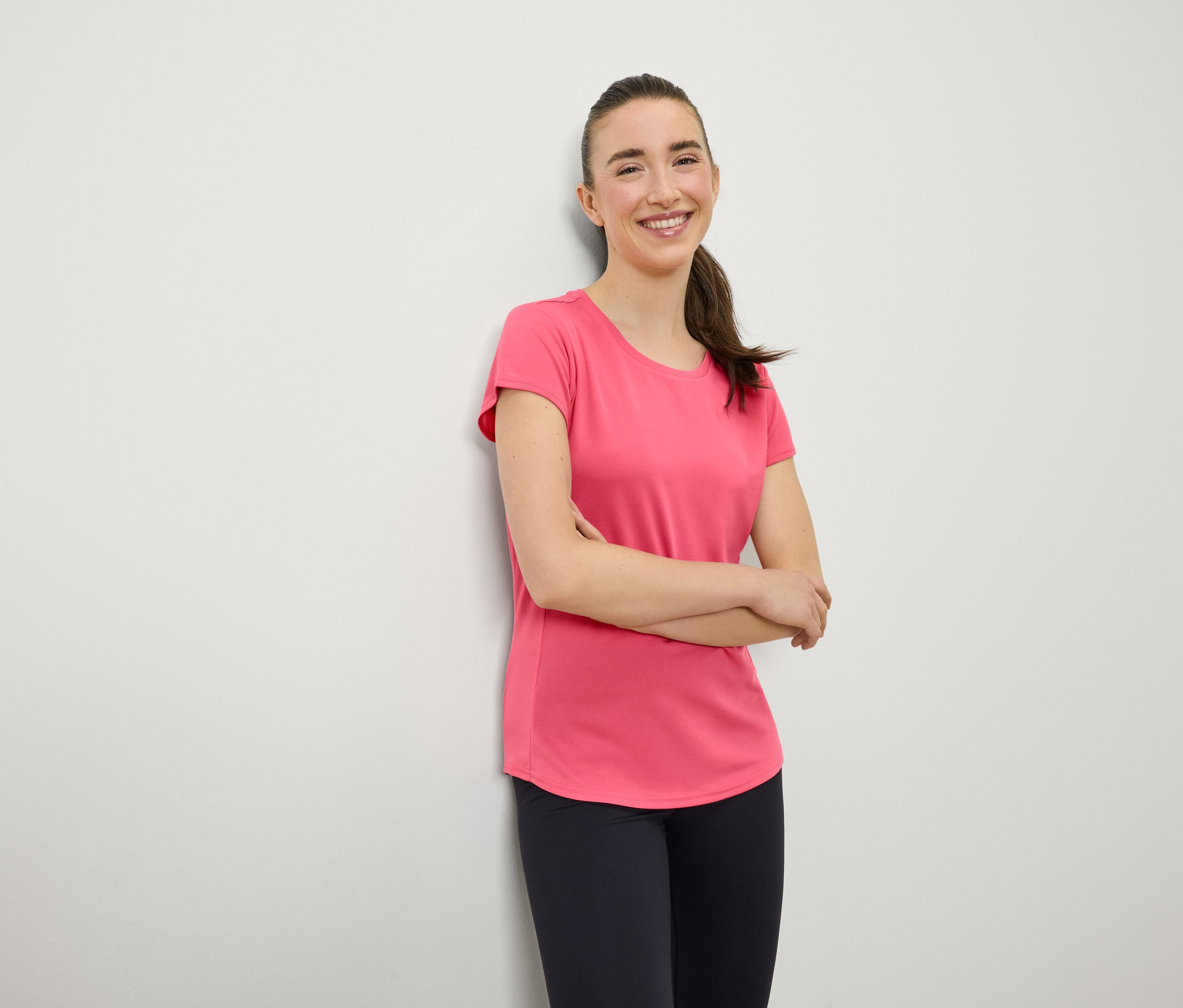 Uśmiechnięta kobieta z brązowymi włosami związanymi w kucyk stoi z założonymi rękami na piersi. Ma na sobie różowy T-shirt i granatowe legginsy.