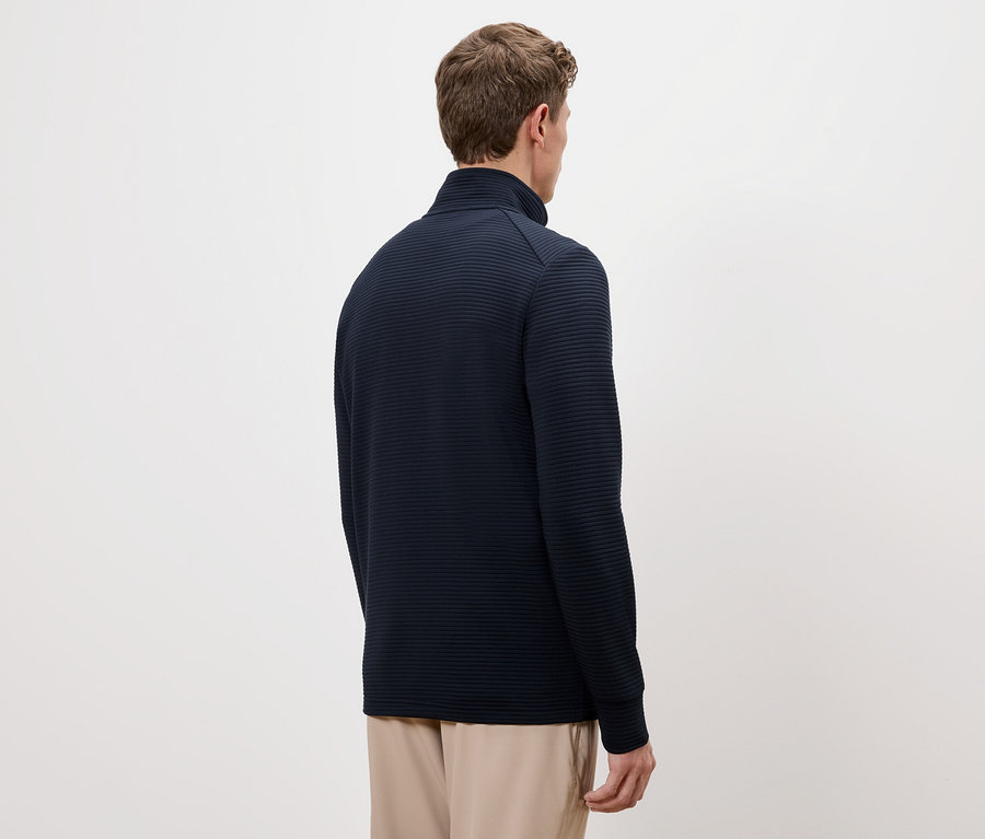 Widok z tyłu mężczyzny z brązowymi włosami w granatowym swetrze ze stójką i beżowych spodniach.