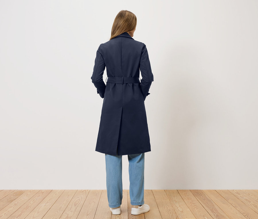 Kobieta stoi tyłem do kamery, ubrana w granatowy trencz, dżinsy i białe buty.