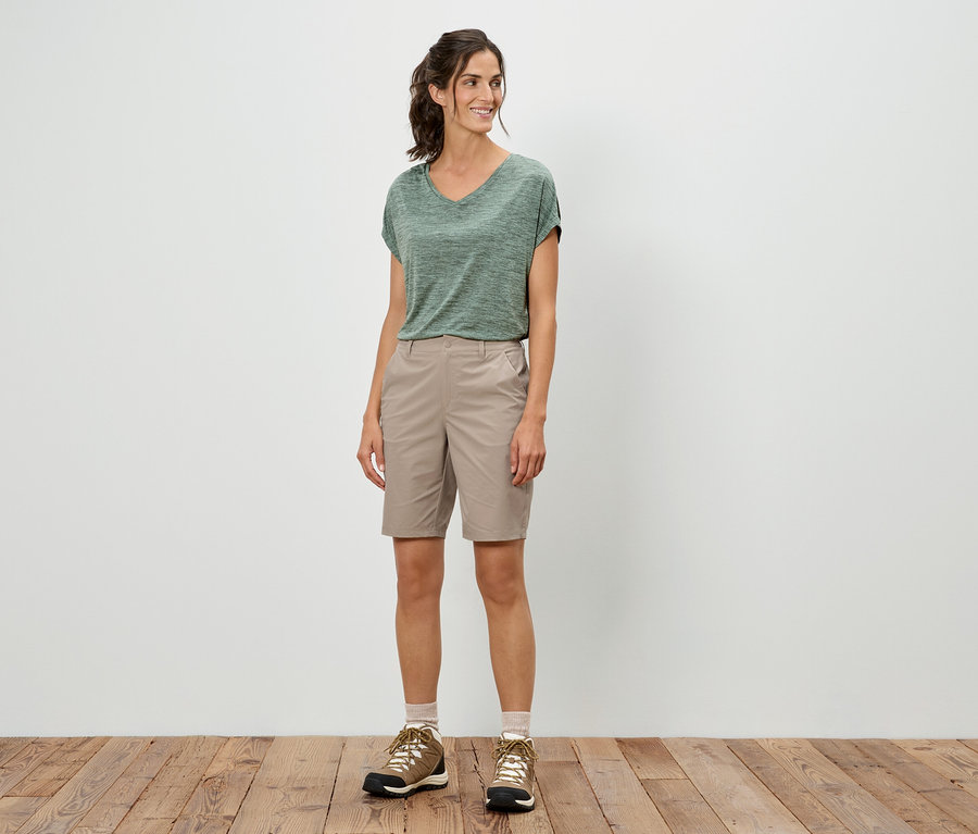Kobieta stoi i ma na sobie zielony T-shirt, szorty i buty trekkingowe.