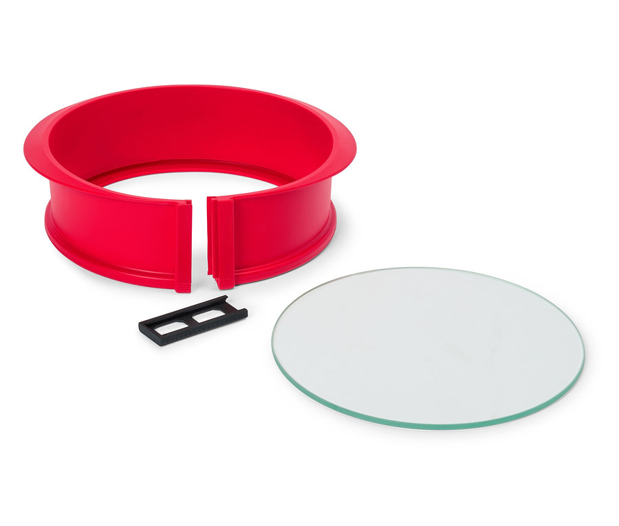 Czerwona, okrągła forma do ciasta z czarnym klipsem i okrągłą szklaną płytą na białym tle.