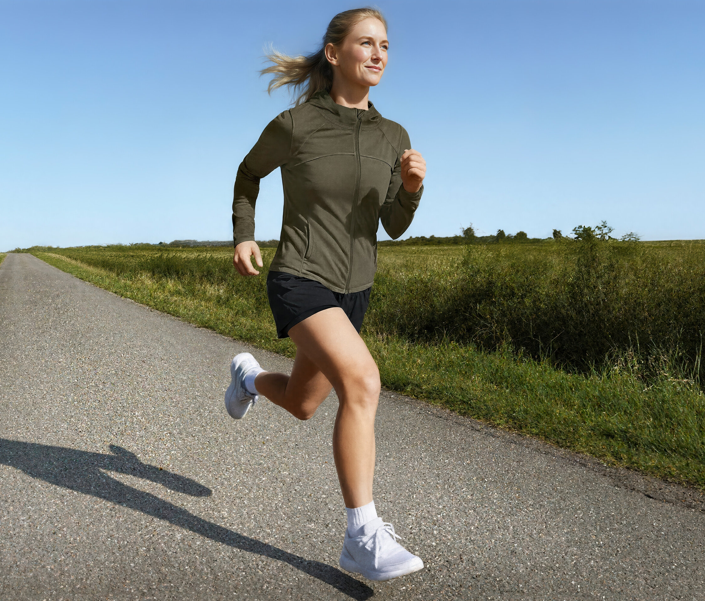 Kobieta biegnie w kurtce sportowej i szortach do biegania 2 w 1 po wiejskiej drodze.