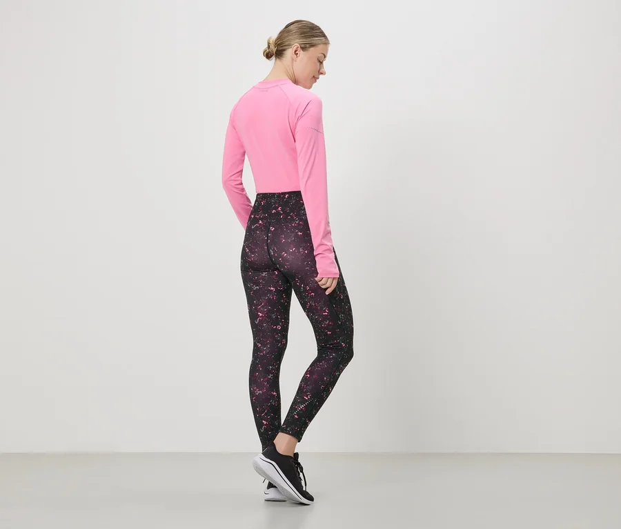Kobieta stoi tyłem, ma na sobie różową bluzkę z długim rękawem, wzorzyste legginsy i czarne buty sportowe.