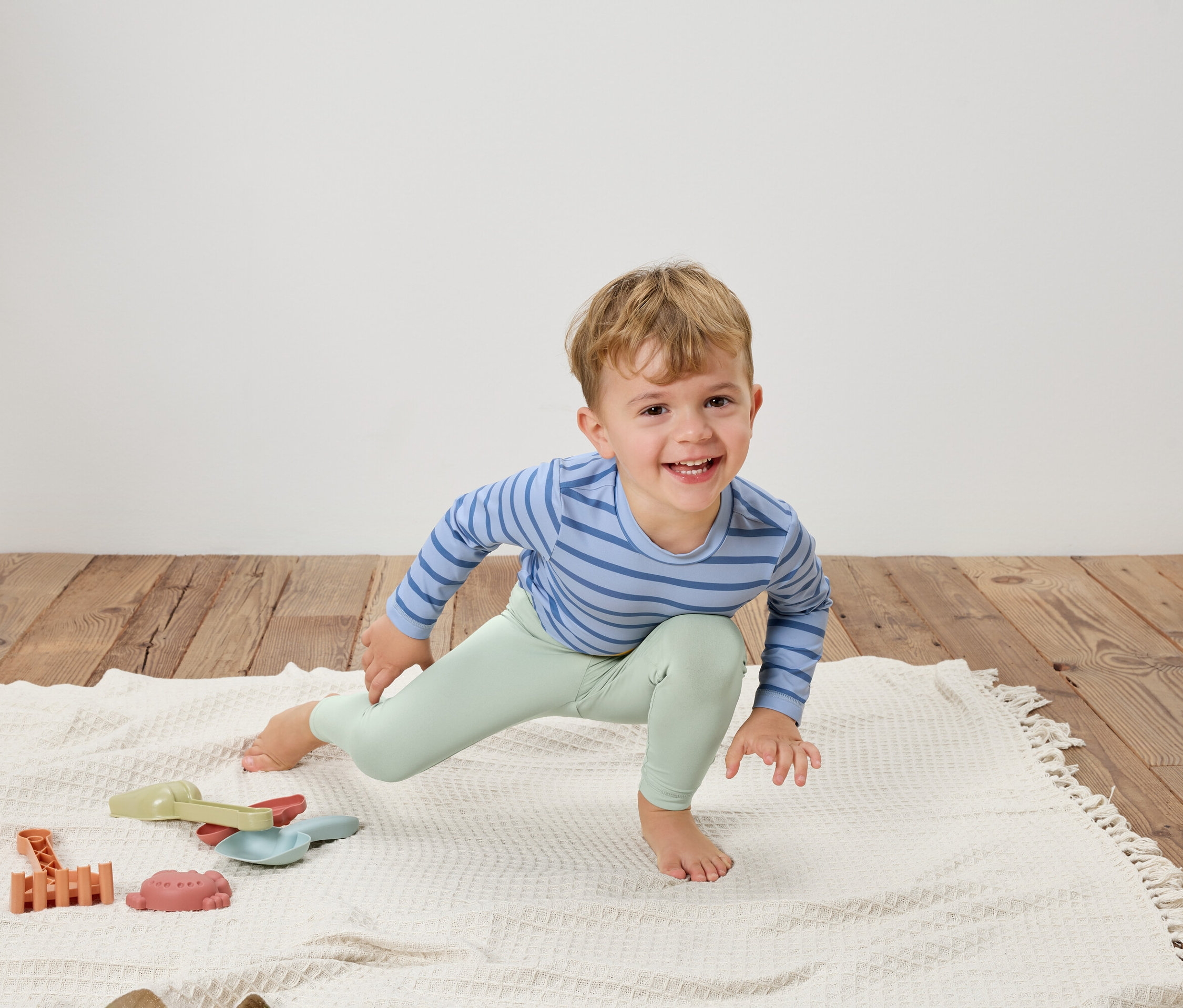 Uśmiechnięty chłopiec w bluzce w niebieskie paski i jasnozielonych legginsach rozciąga się na białym kocu obok zabawek.