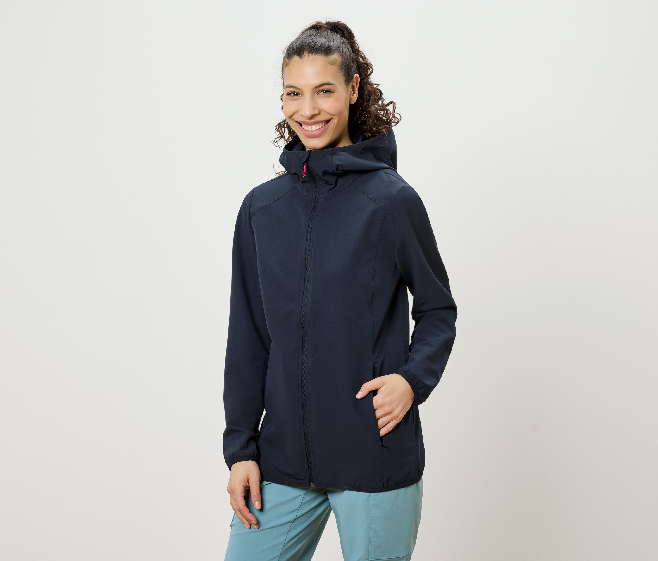 Uśmiechnięta kobieta z kręconymi włosami upiętymi w kok, ubrana w granatową kurtkę z kapturem i niebieskie spodnie, z ręką w kieszeni kurtki.