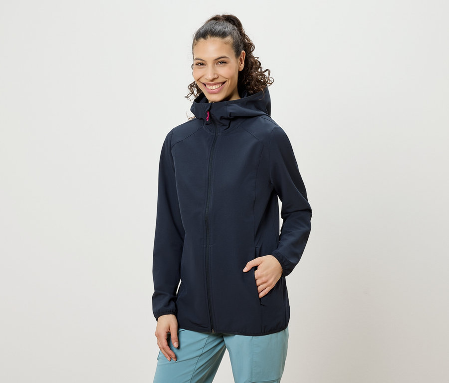 Uśmiechnięta kobieta z kręconymi włosami upiętymi w kok, ubrana w granatową kurtkę z kapturem i niebieskie spodnie, z ręką w kieszeni kurtki.