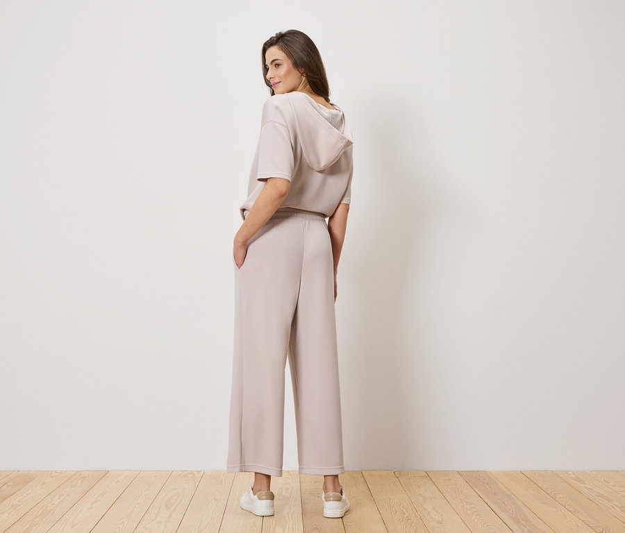 Kobieta z brązowymi włosami, sfotografowana od tyłu, ma na sobie jasnobrązową bluzę z kapturem i szerokie spodnie.