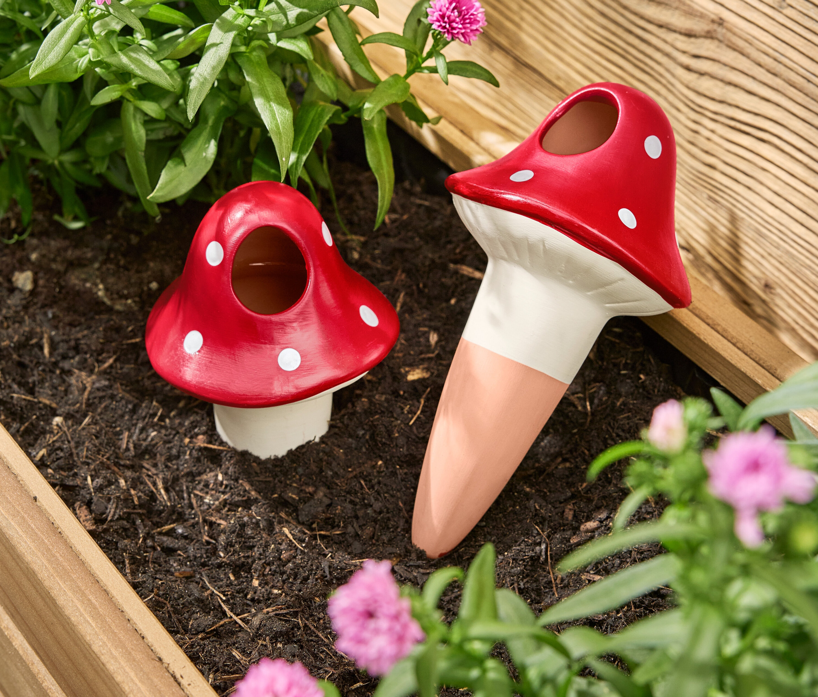 Dwie czerwone kulki nawadniające w kształcie grzybów z białymi kropkami stoją w skrzynce na kwiaty pomiędzy kwiatami i roślinami.