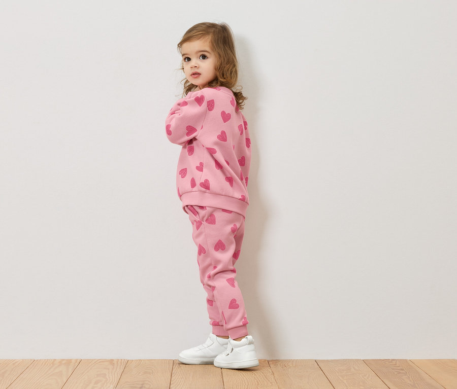 Mała dziewczynka stoi bokiem, ubrana w różową bluzę i spodnie w serduszka, do tego białe buty.