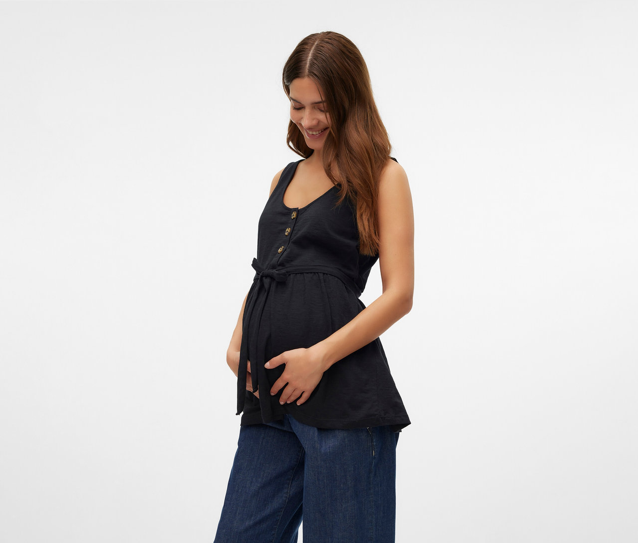 Kobieta trzyma się za brzuch, ma na sobie czarny top do karmienia piersią MAMALICIOUS „MLEVI LIA” i szerokie dżinsy.